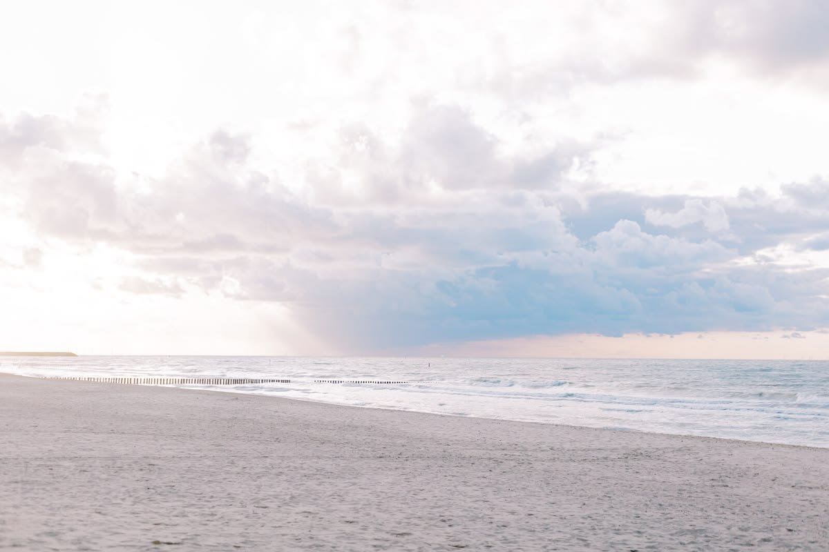 A calm beach with light-colored sand, gentle waves, a distant shoreline, and cloudy sky with patches of sunlight.