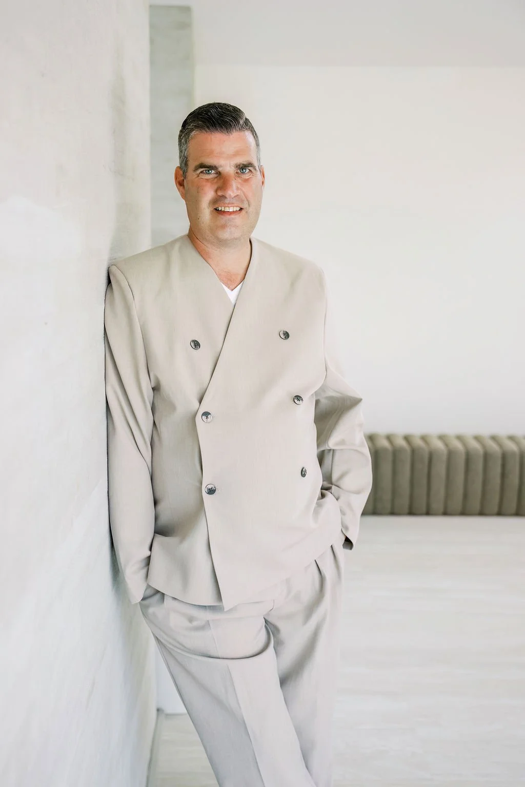 A man in a beige double-breasted chef's coat standing and smiling against a white wall.