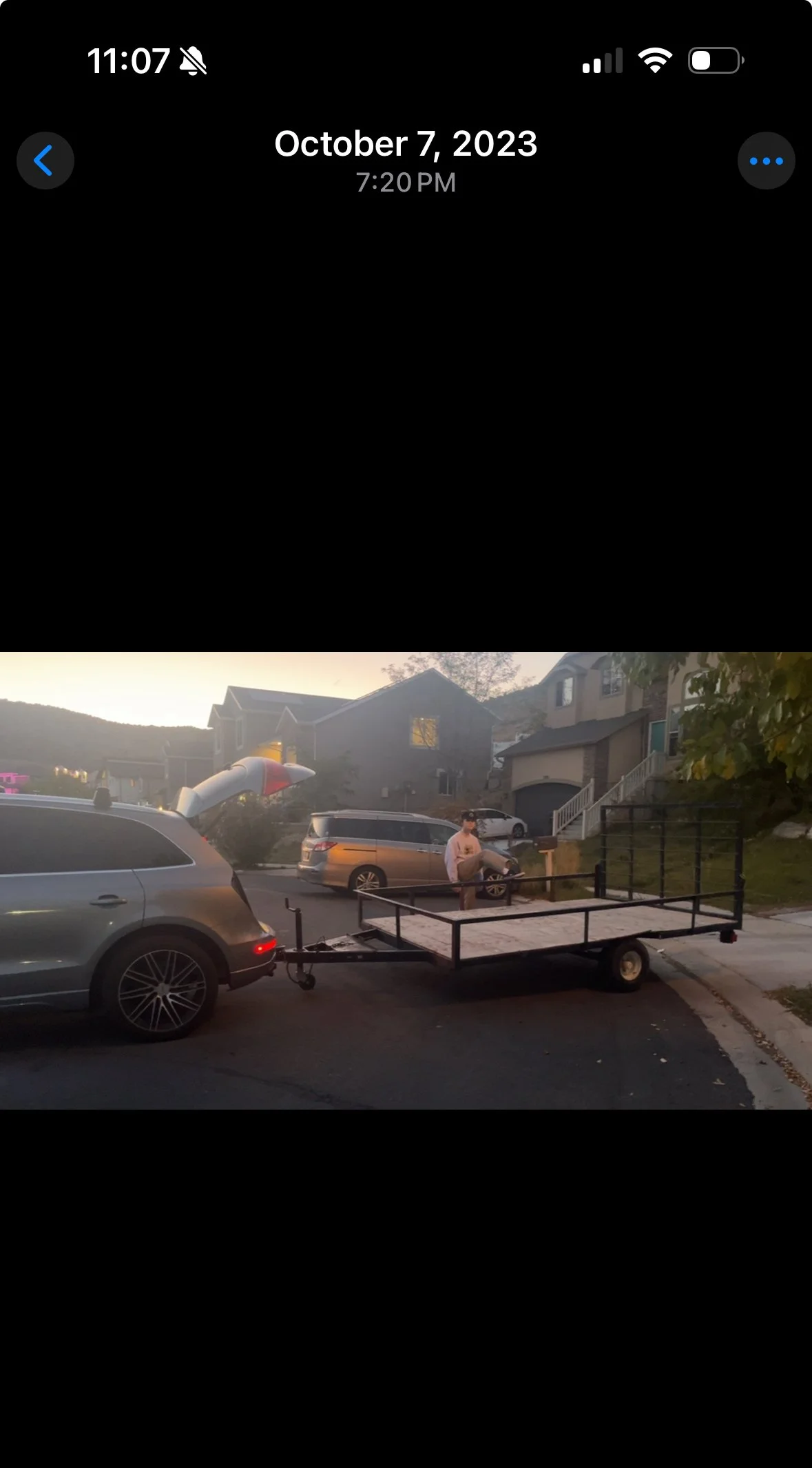 A man sitting on a small vehicle with loaded trailers parked in a residential neighborhood with houses and cars in the background during sunset.
