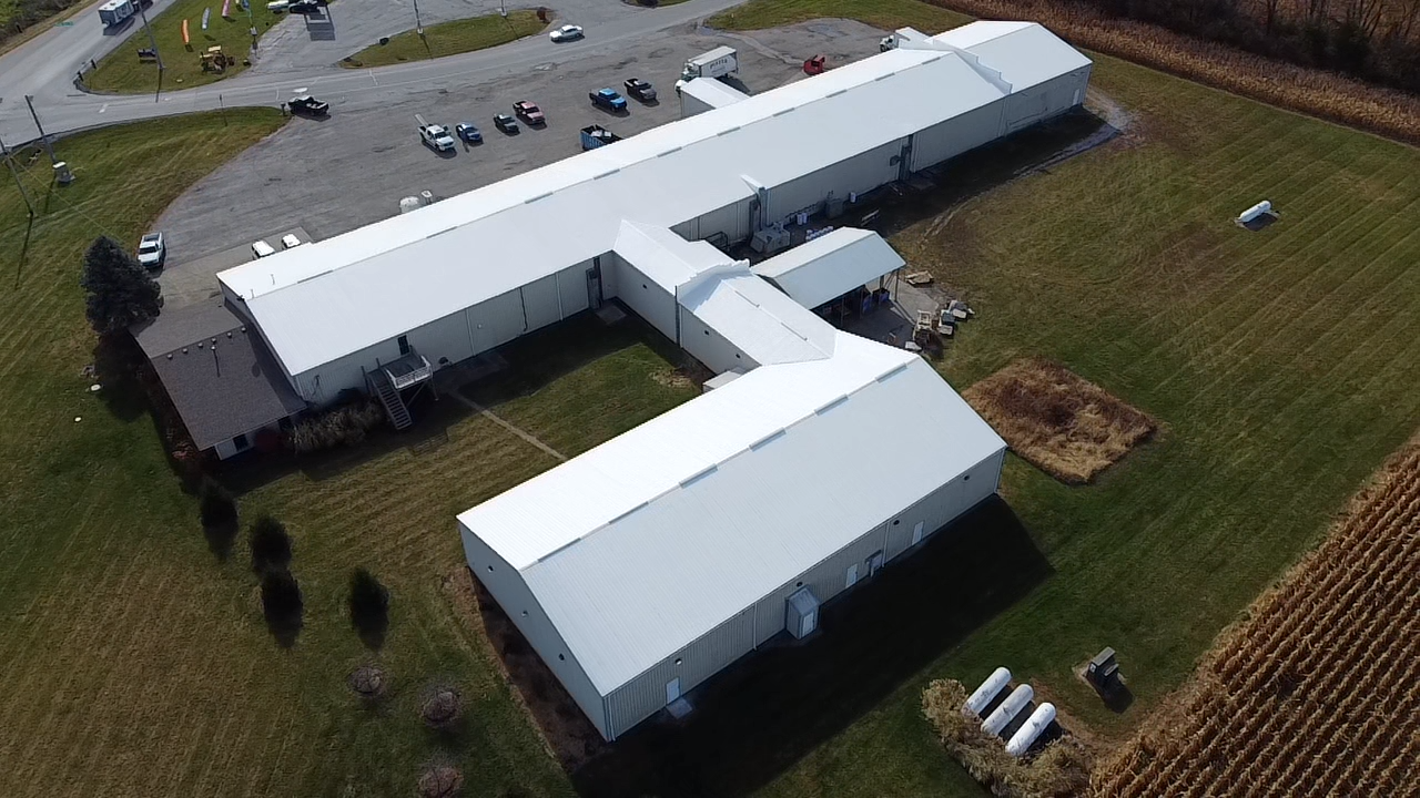 Aerial view of a large white industrial or agricultural building complex with metal roofing, surrounded by grassy fields, parked cars, and a small residential house nearby.