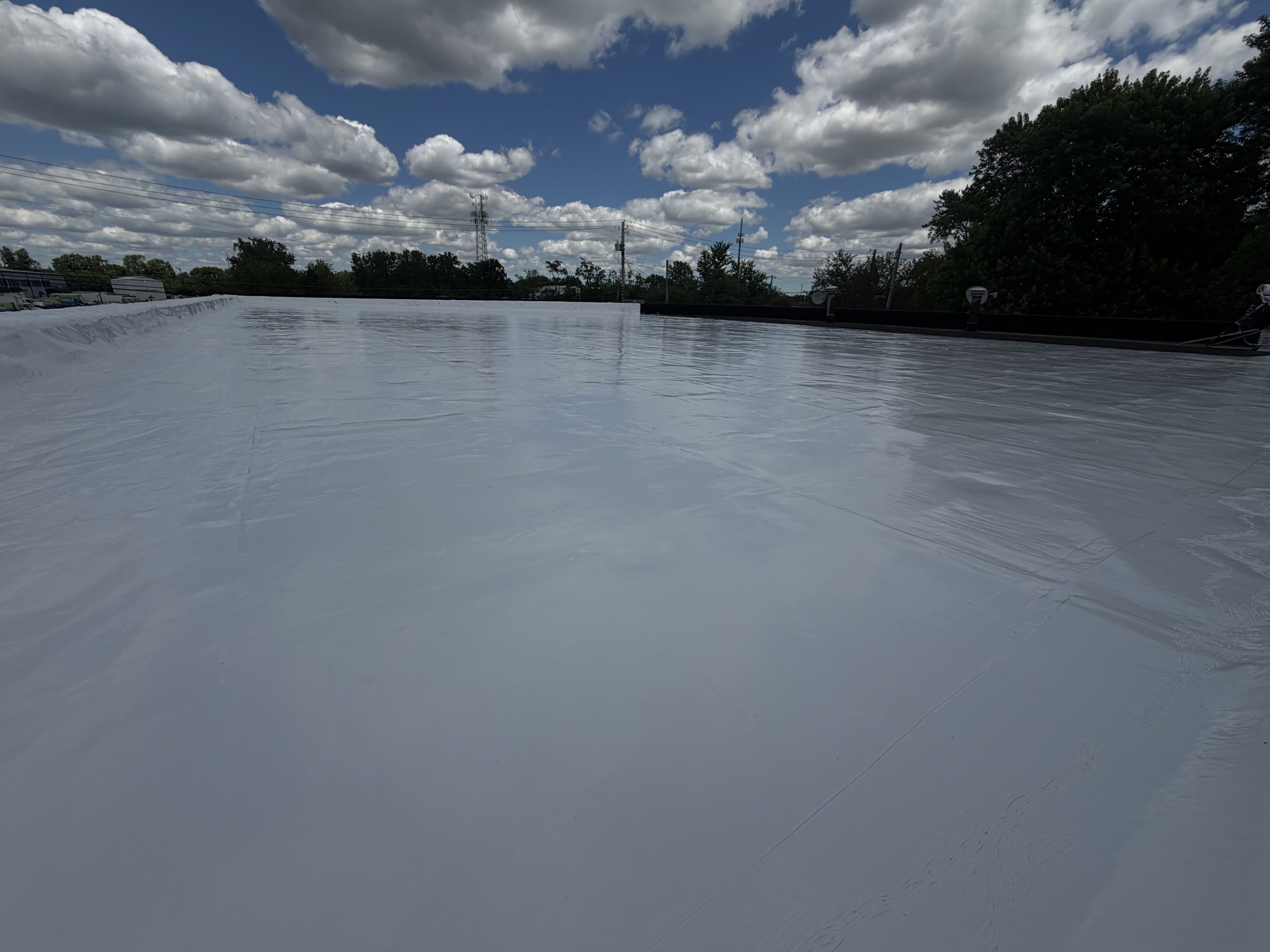 A flat rooftop covered with a white reflective barrier, with trees and power lines in the background under a blue sky with scattered fluffy clouds.
