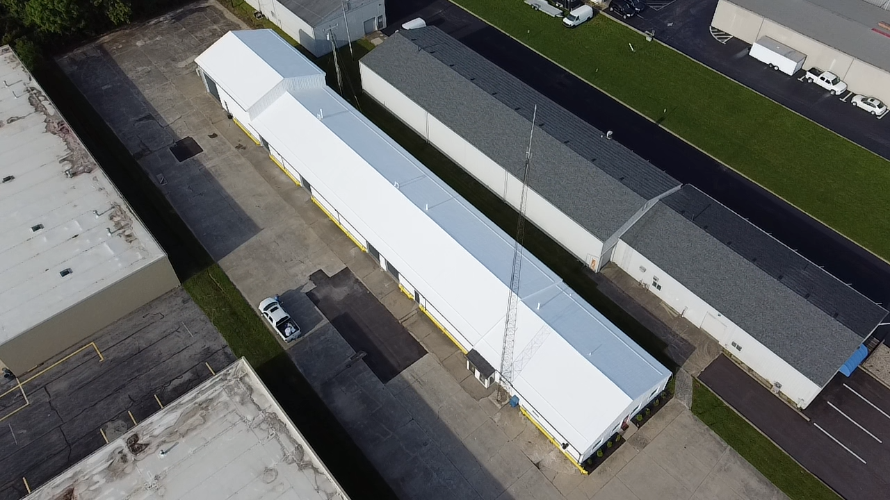 Aerial view of a white, elongated portable building with a curved roof, parked next to other industrial buildings and structures in an industrial area.