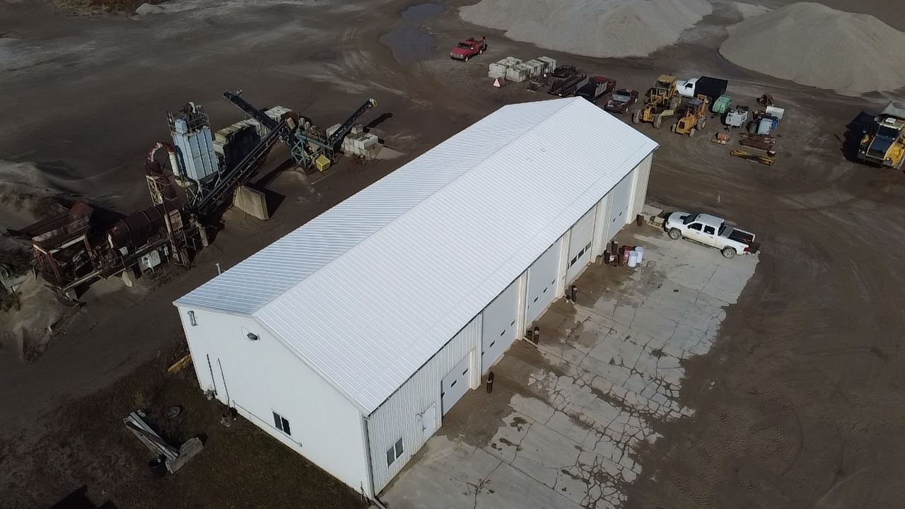 Aerial view of a large white industrial building, surrounded by parked vehicles, construction equipment, and a dirt lot with machinery and scattered materials.