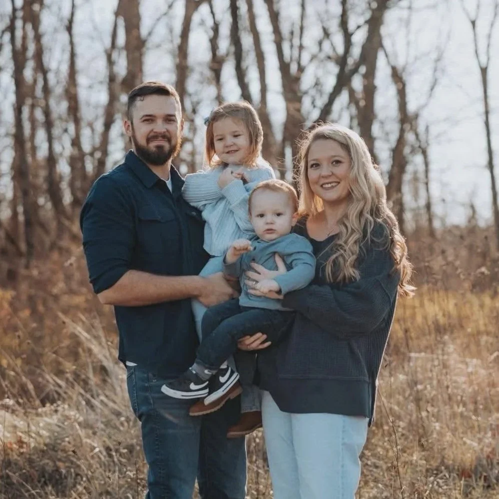 A family of four standing outdoors in a wooded area, with the father holding a young girl and the mother holding a toddler, all dressed casually for fall.
