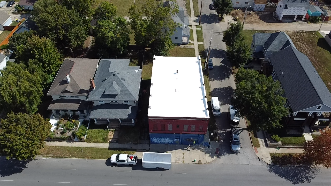 Aerial view of a city block showing two residential houses and one commercial building with a white roof, on a street lined with trees, parked vehicles, and sidewalks.