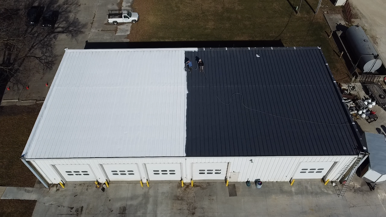 An aerial view of a large building with a metal roof, partially painted black on one side and white on the other, with two workers standing on the roof.
