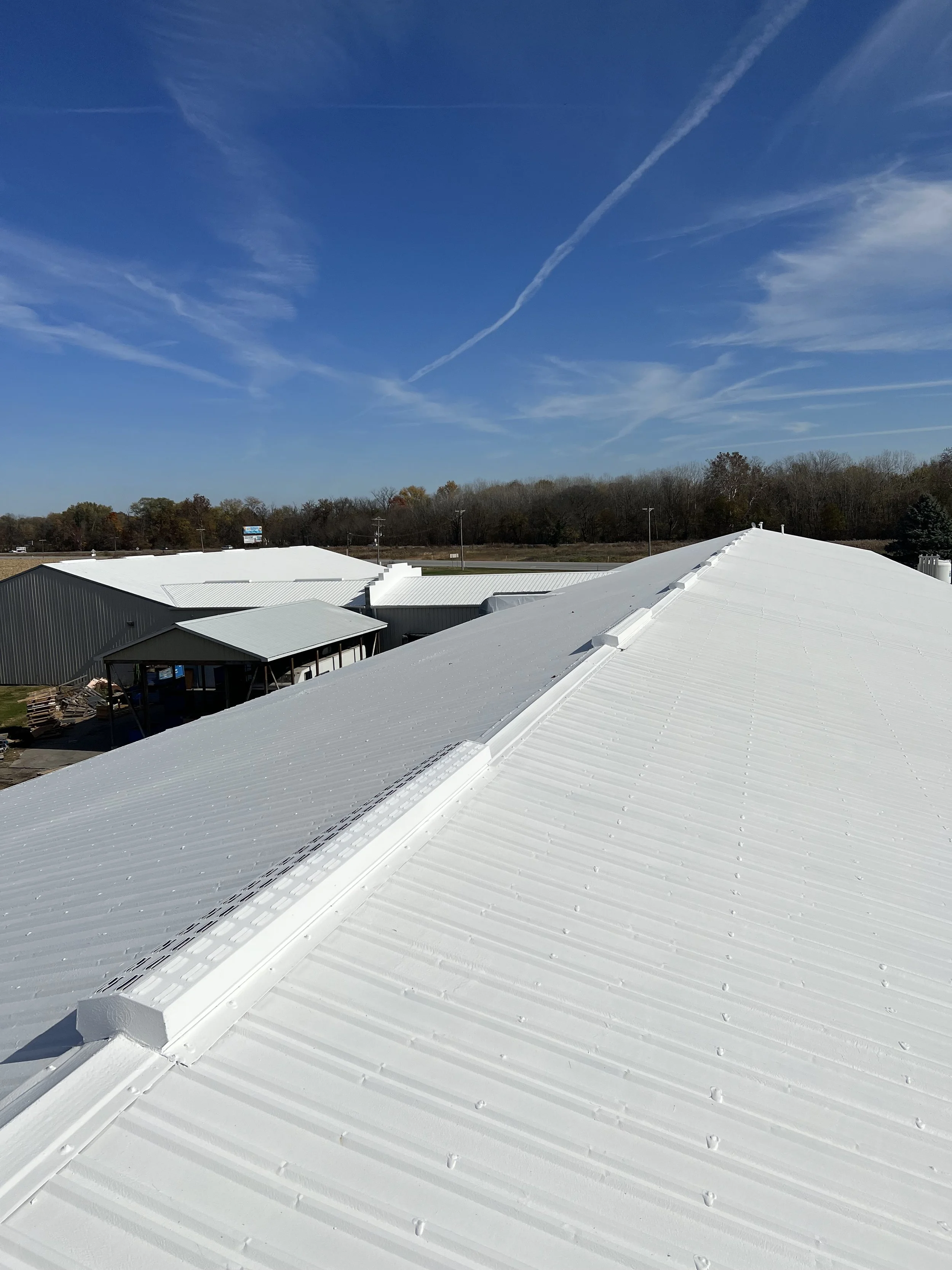 White metal rooftops of industrial buildings under a blue sky with wispy clouds and contrails.
