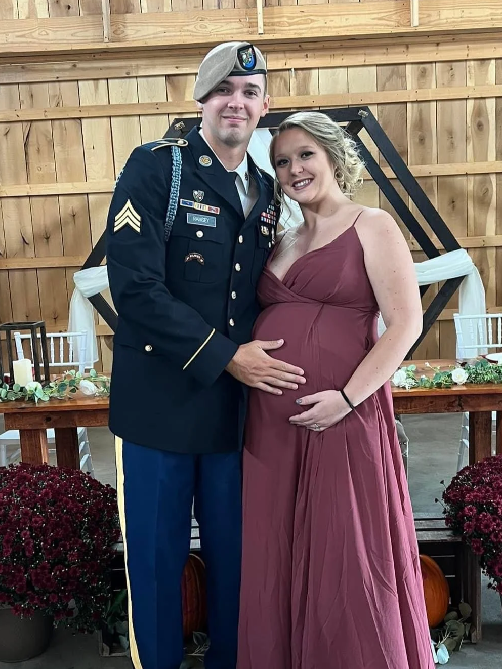 A pregnant woman and a man in military uniform standing together indoors, smiling at the camera, with wooden decor and a table with candles and flowers behind them.