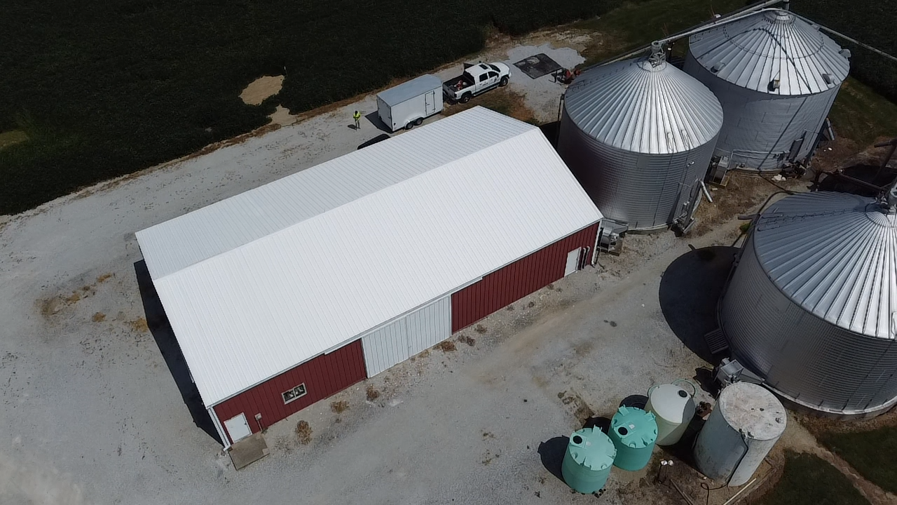 Aerial view of a farm setup including a long red and white barn, three large metal grain silos, several green and white storage tanks, parked vehicles, and a person walking nearby.