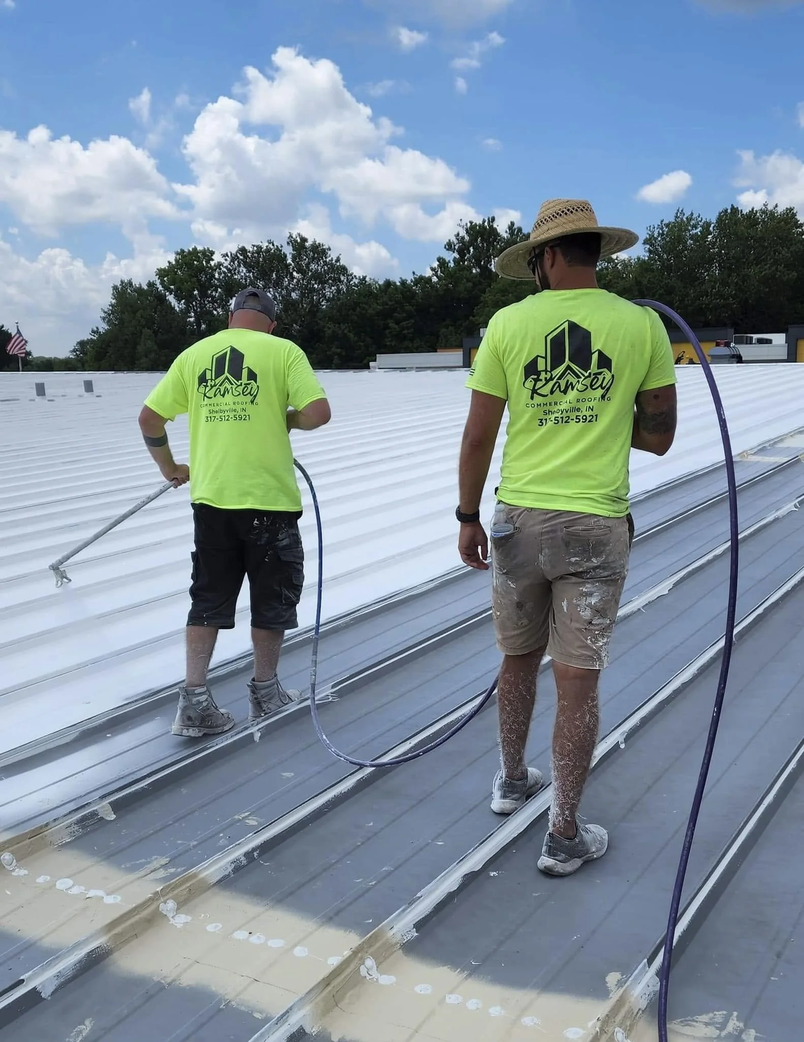 Two workers in bright green shirts and shorts are cleaning or applying a coating on a large white metal roof. One worker is wearing a wide-brimmed hat, and they are using hoses or spray equipment. A landscaped background with trees and a blue sky with scattered clouds is visible.