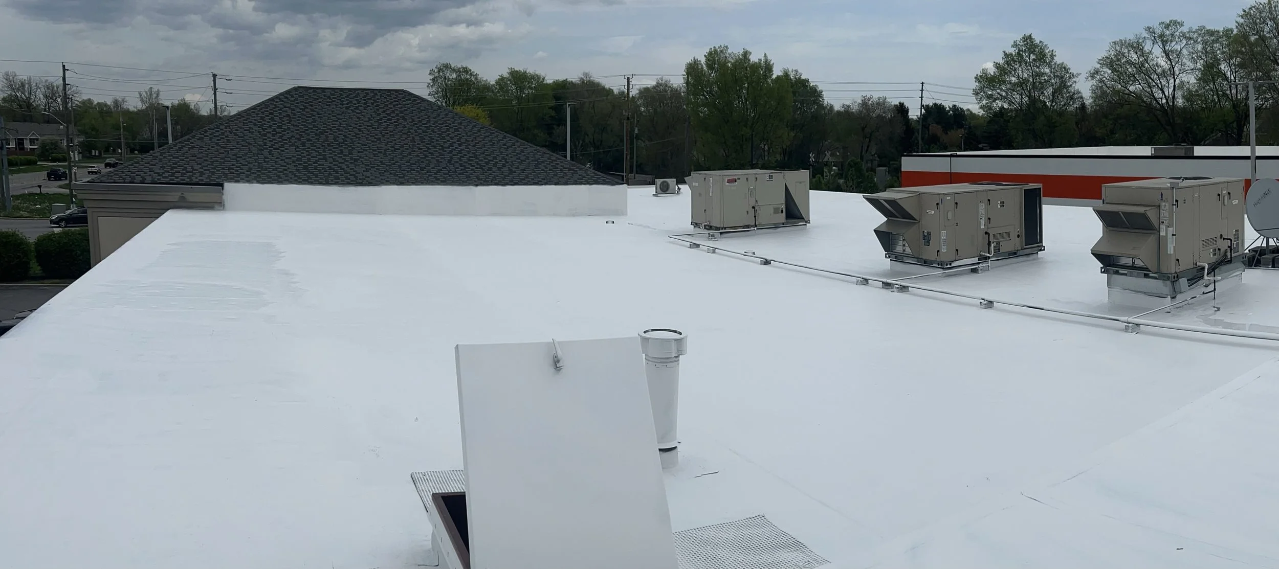 View of a flat white commercial building roof with three HVAC units and a vent pipe, with a background of trees, power lines, and cloudy sky.