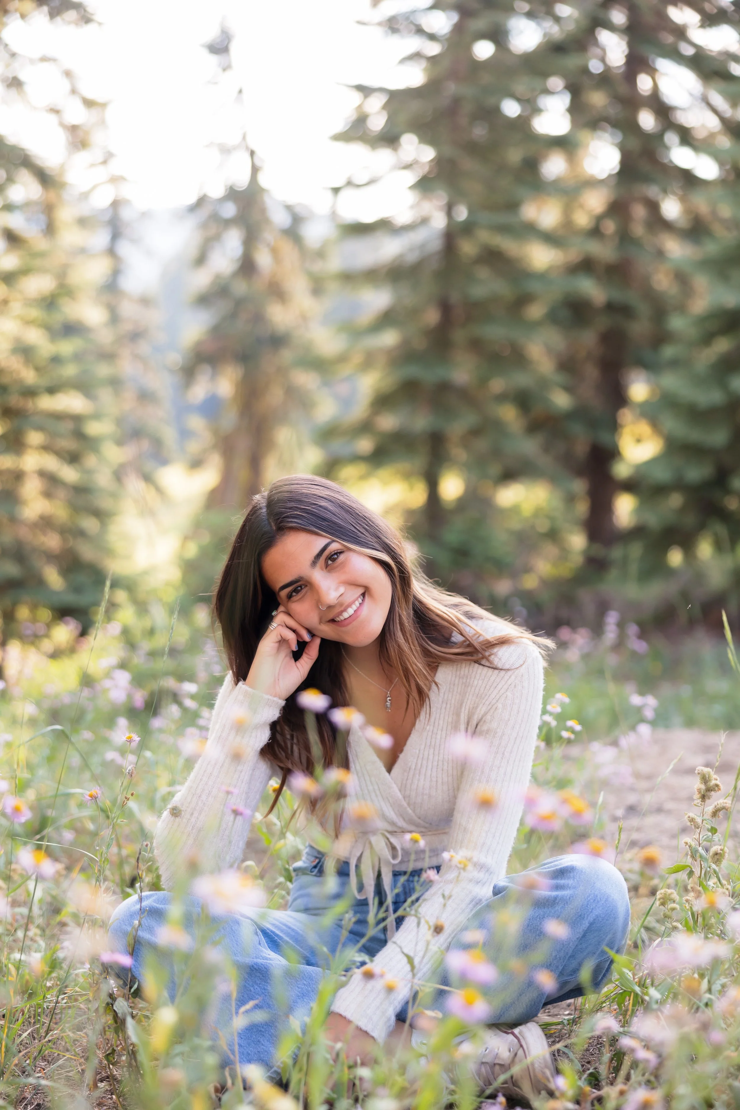 A young woman with long brown hair and a nose ring smiling while sitting in a field of wildflowers, with tall trees and sunlight in the background.