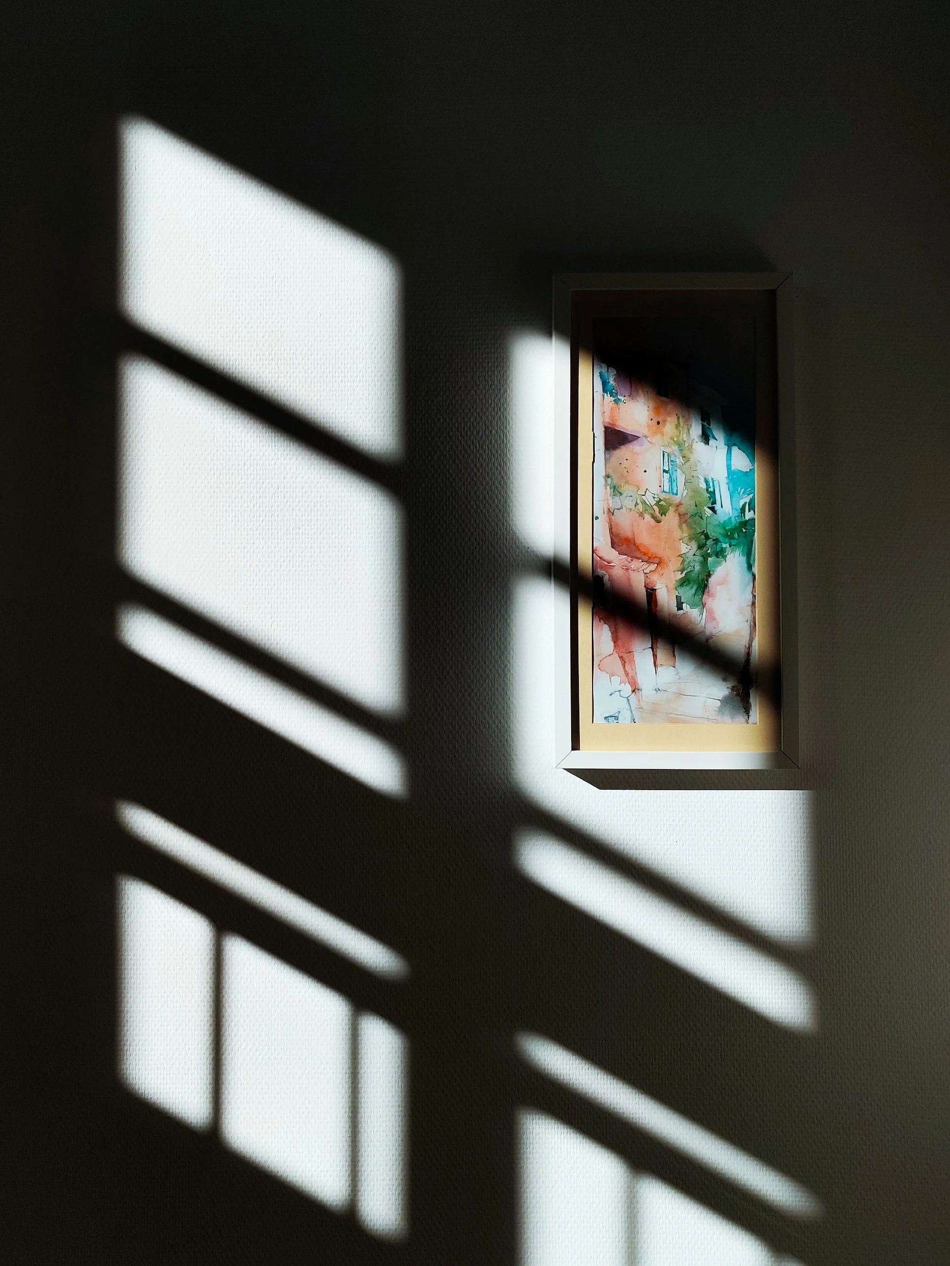 Shadow of a ladder cast on a wall next to a framed watercolor painting of a landscape.