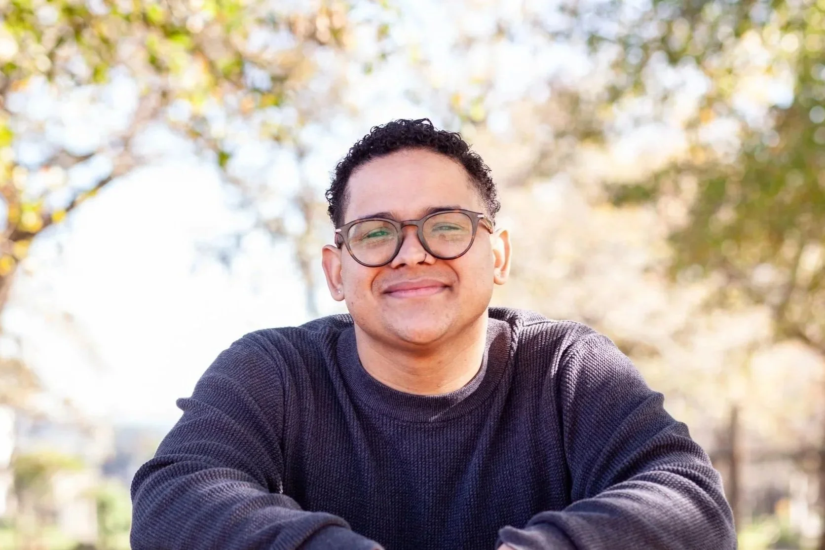 A young man with glasses smiling while taking a selfie outdoors with trees in the background.