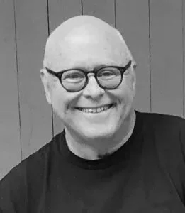 A smiling man with glasses and a bald head, wearing a black shirt, in front of a wood-paneled background.