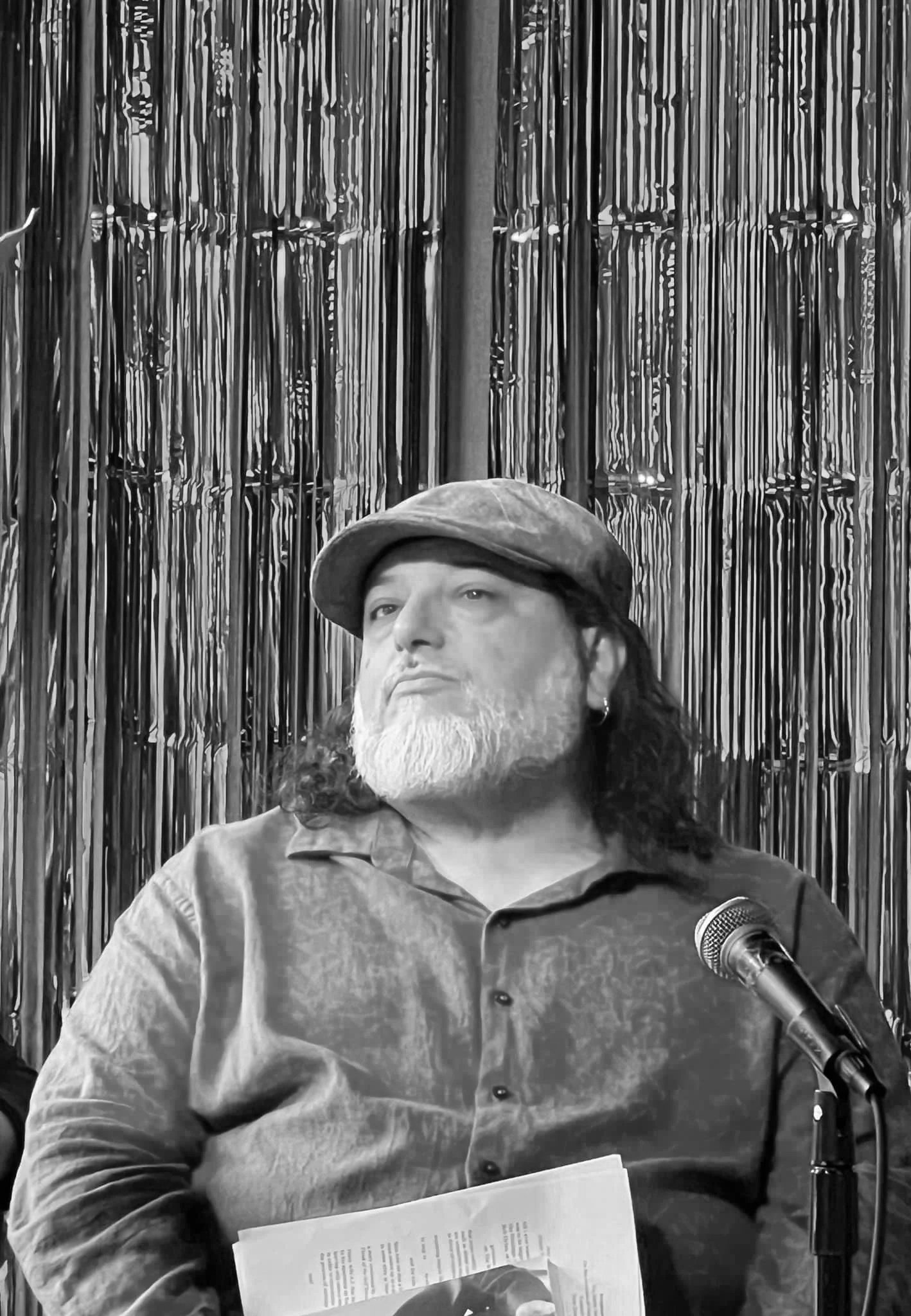 A man with long hair, a beard, and wearing a cap and a button-down shirt, is sitting in front of a curtain made of hanging metallic strips. He is looking slightly upward, and there is a microphone in front of him, with some papers on his lap.