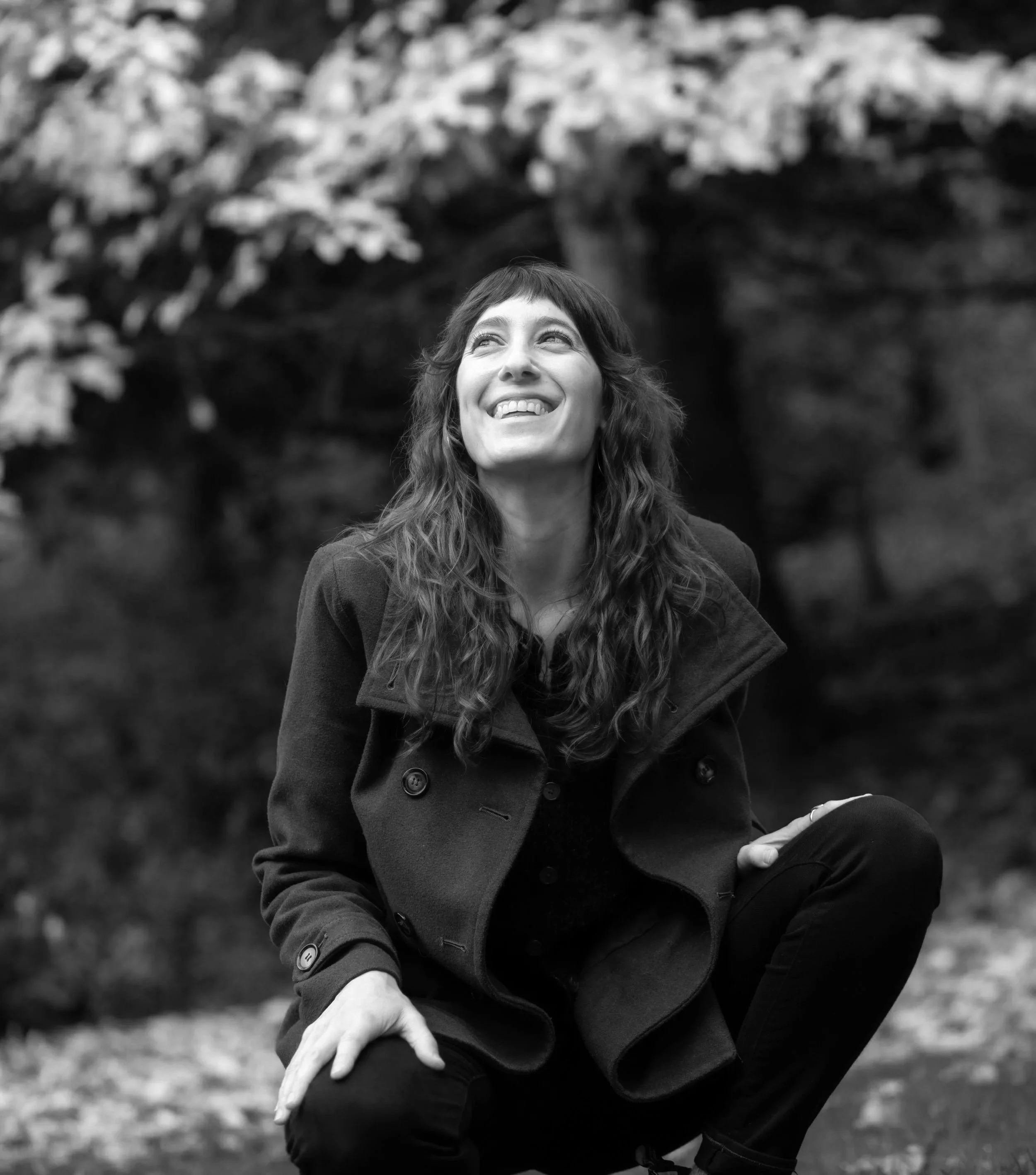 Black and white photo of a smiling woman with long wavy hair, sitting outdoors wearing a coat, with blurred trees in the background.