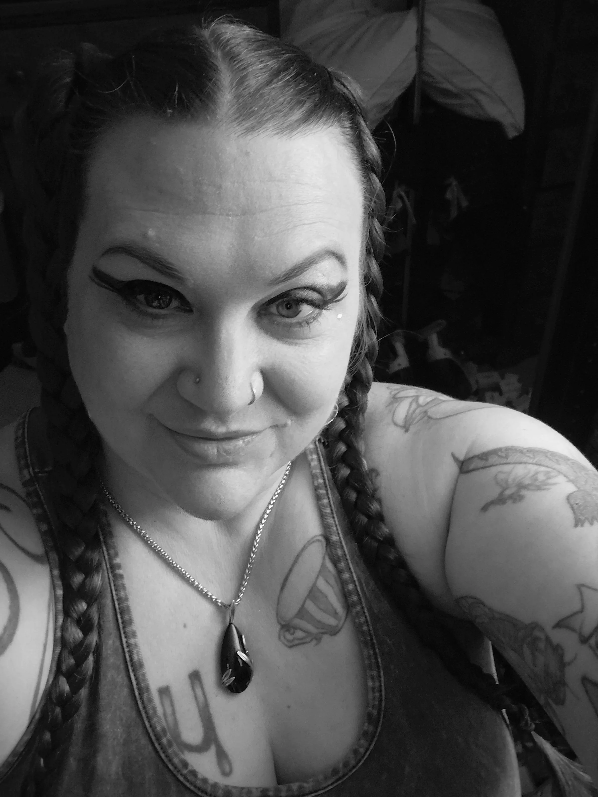 A black and white photo of a woman with braided hair, tattoos on her arm, a nose piercing, and wearing a necklace with a large black pendant. She is smiling and looking at the camera.