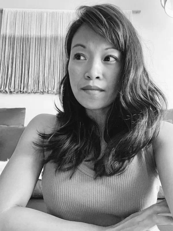 Black and white photo of a woman with shoulder-length wavy hair sitting indoors, looking off to the side, wearing a sleeveless top.