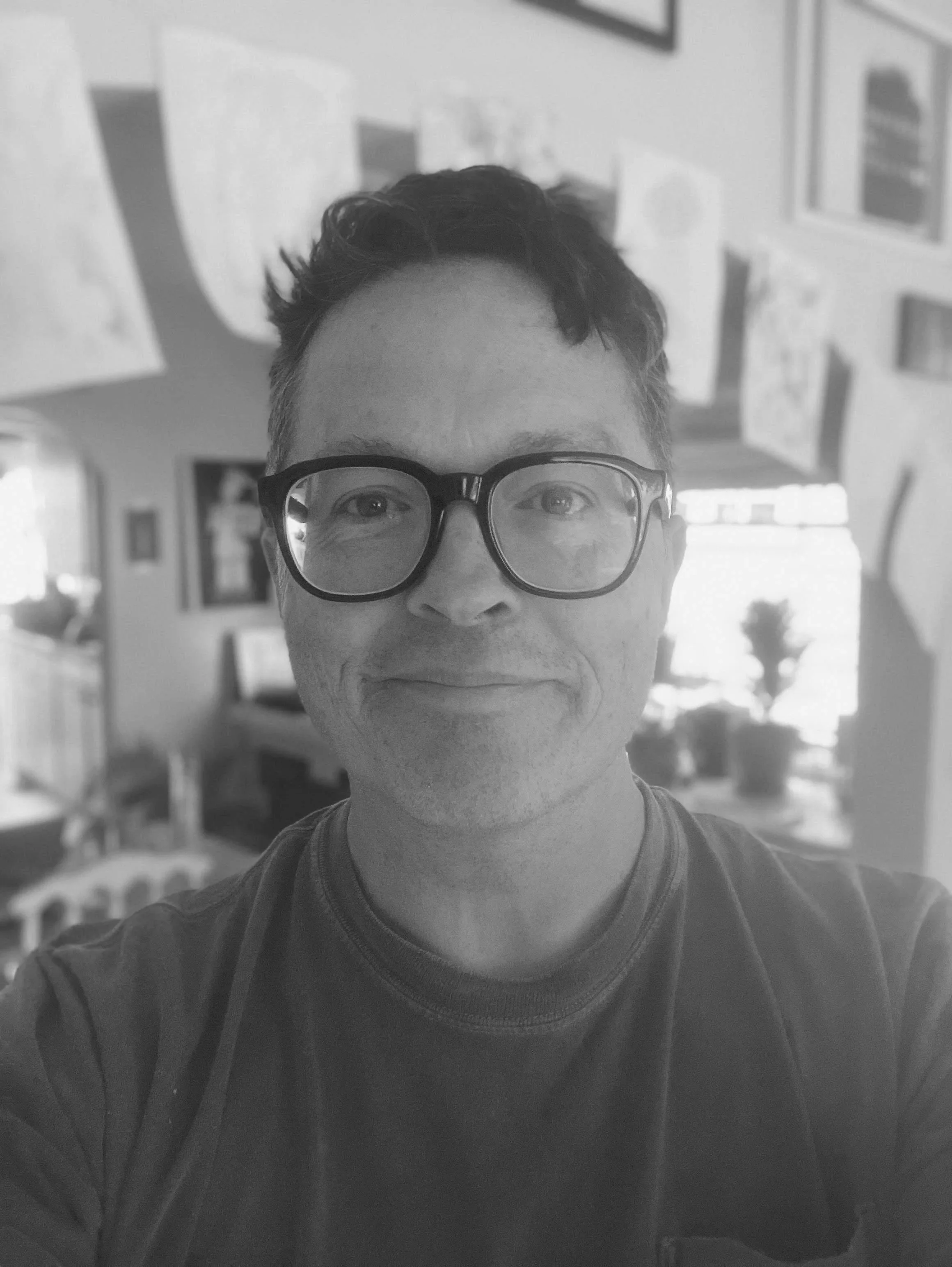 A black-and-white photo of a young man wearing glasses with a partial view of his face, short curly hair, and a casual T-shirt, standing inside a room with artwork on the walls.