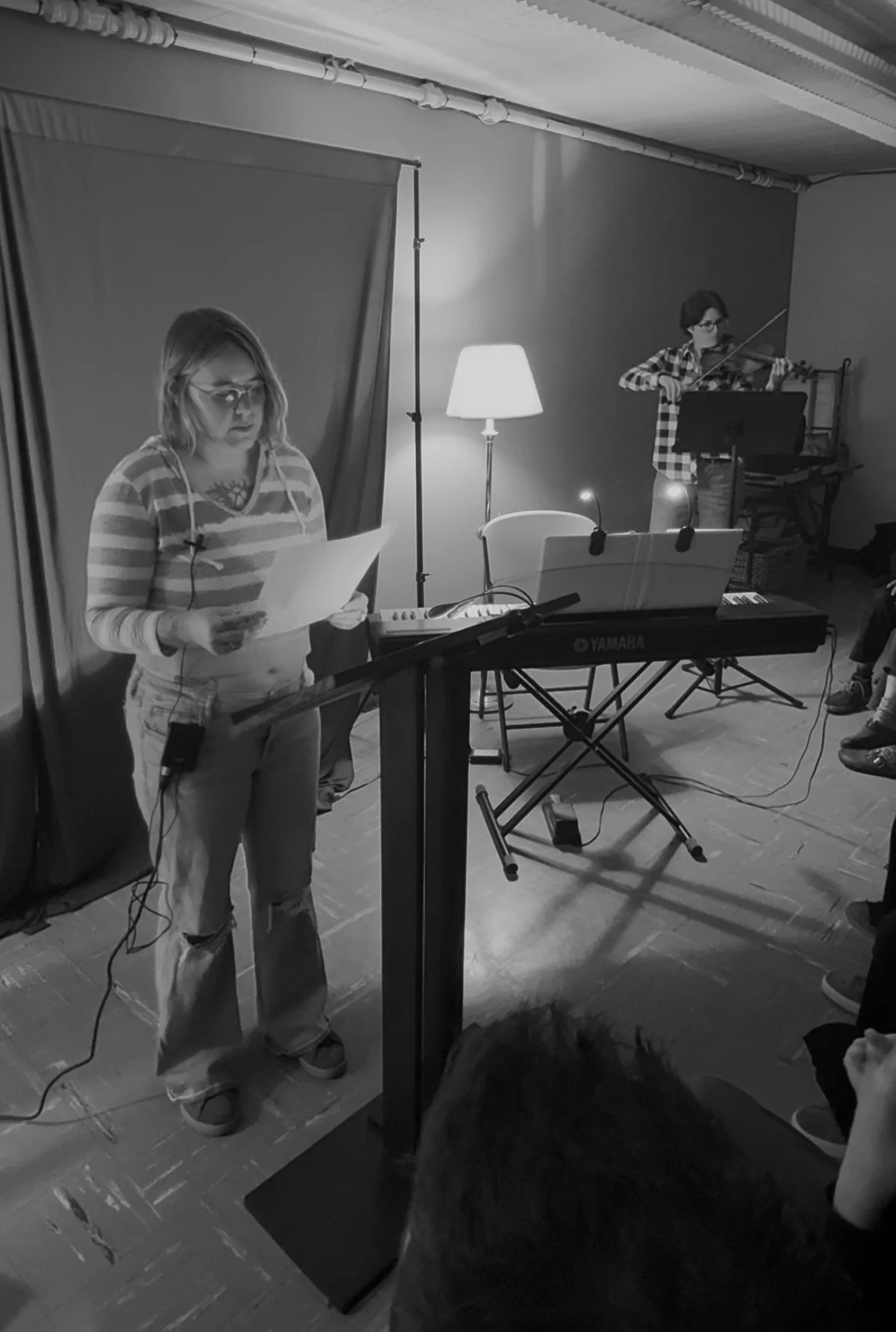 A woman with glasses and a striped shirt is reading from a paper in front of a music stand. A keyboard and a person playing the violin are visible in the background.