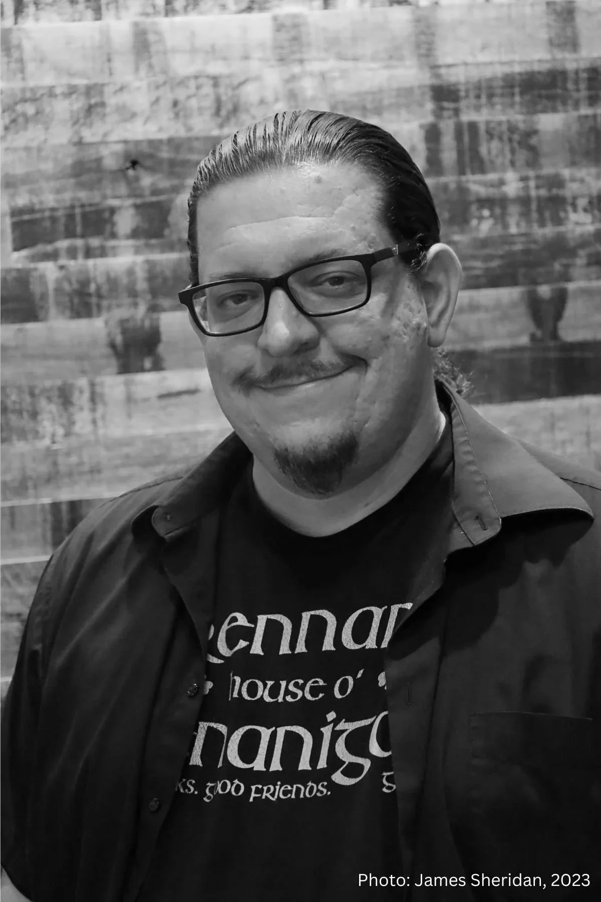 A black and white photo of a man with glasses, a mustache, and a goatee, smiling while standing against a wooden background. He is wearing a dark shirt with partially visible light-colored text and a dark jacket.