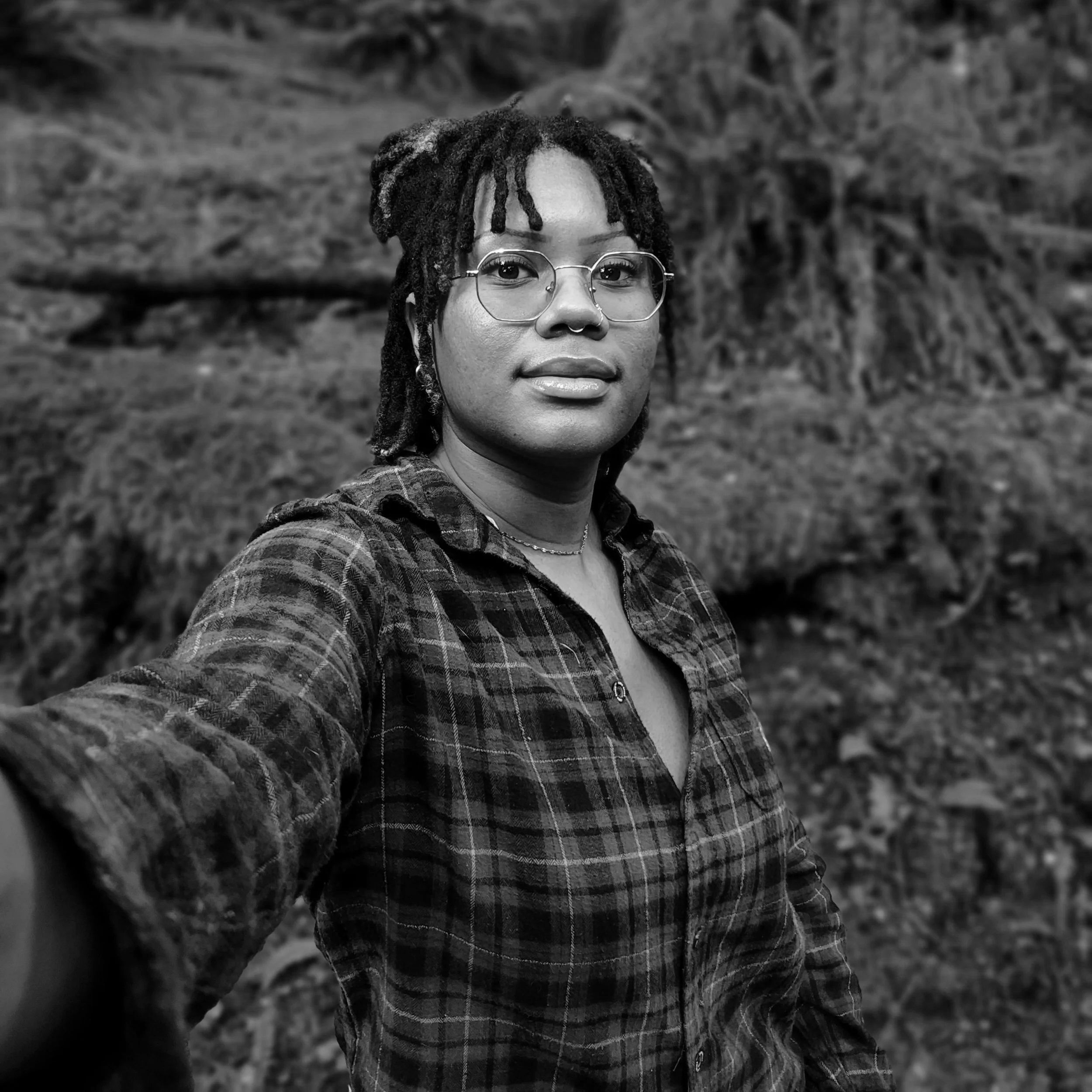 A black and white portrait of a woman with glasses and dreadlocks, outdoors with a blurred natural background.