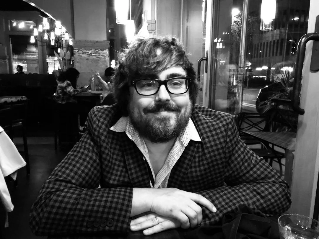 A man with glasses, a beard, and messy hair sitting at a table inside a restaurant, smiling at the camera.
