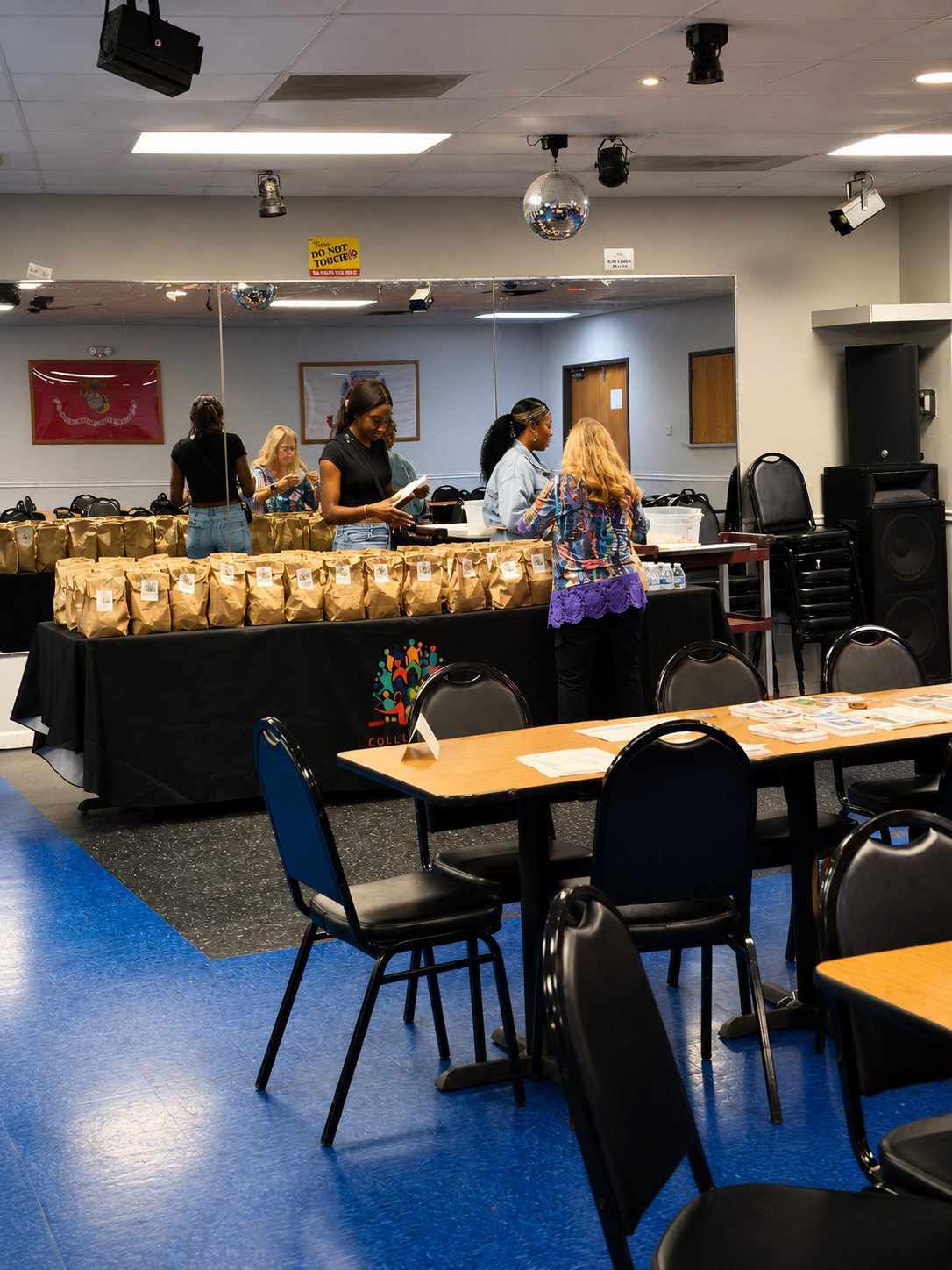 People preparing meal bags at a community event with tables and stacked chairs in a multi-purpose room.