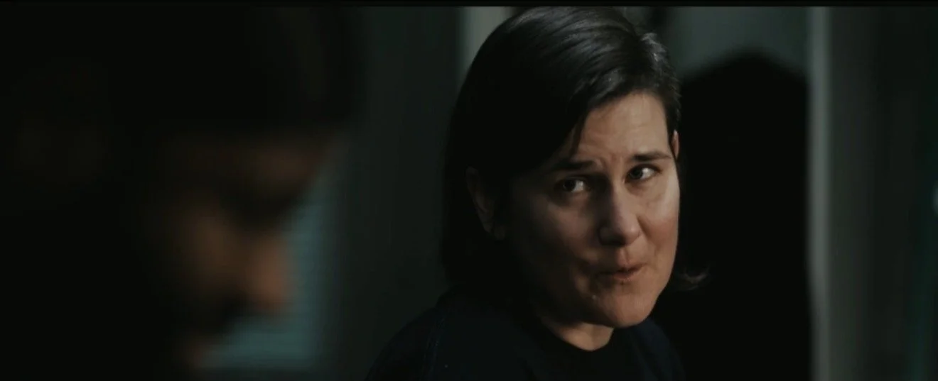 A woman with short dark hair making a skeptical or suspicious facial expression in a dimly lit room.