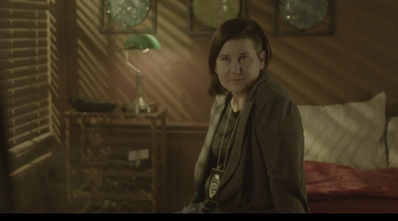 A woman with short brown hair, wearing a dark blazer and a badge, sitting on a bed in a room with wooden walls, a green desk lamp, and window blinds.