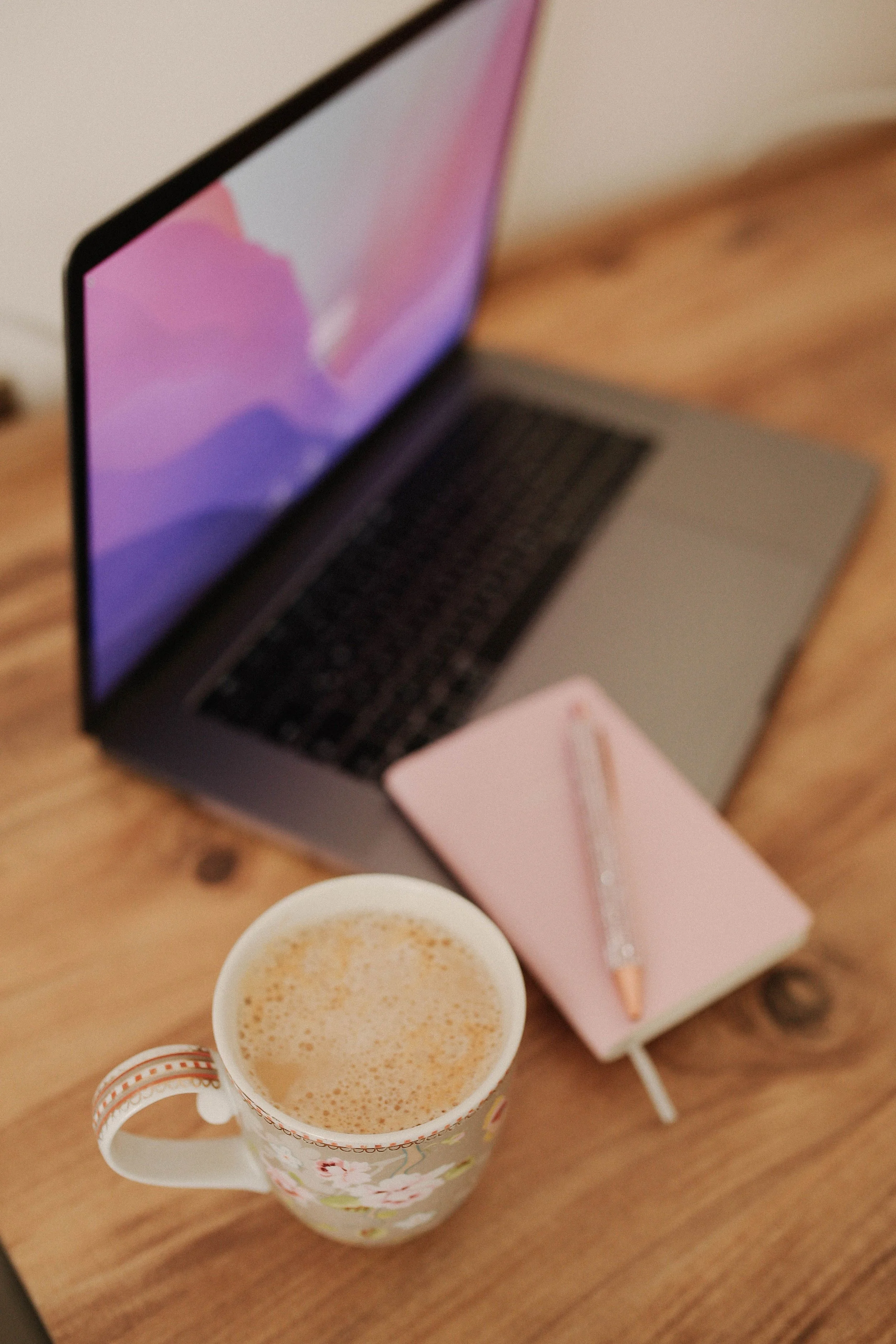 A floral decorated mug filled with a frothy coffee beverage, placed on a wooden table along with a pink notebook, a pen, and a pink box, with a laptop in the background.