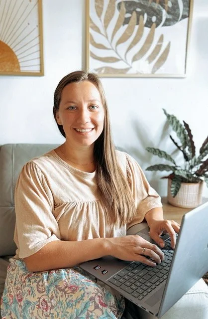 A woman sitting on a sofa using a laptop, smiling at the camera, with framed artwork and a plant in the background.