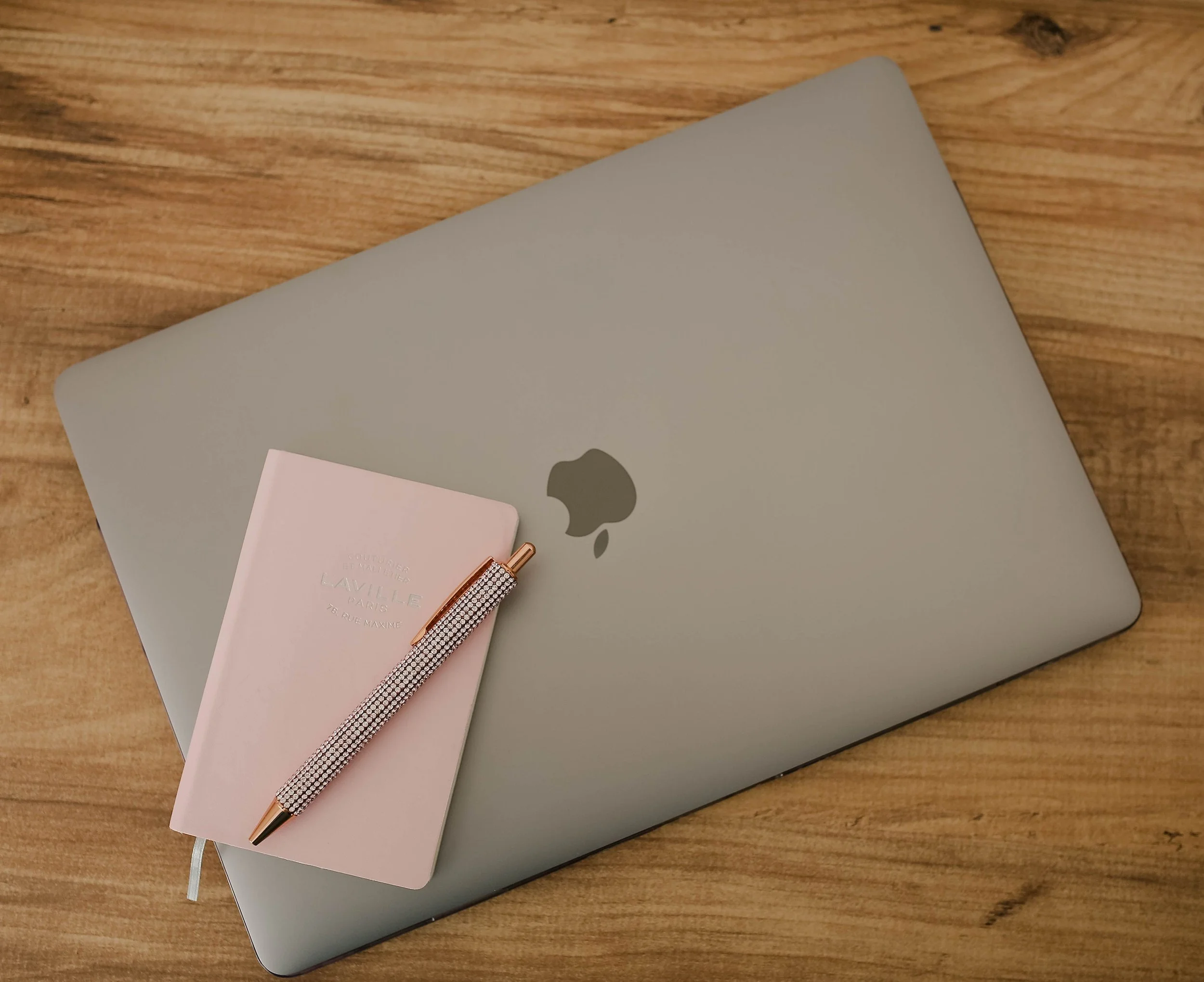 A MacBook laptop with a gray cover on a wooden surface, a pale pink notepad, and a rose gold pen with rhinestones.