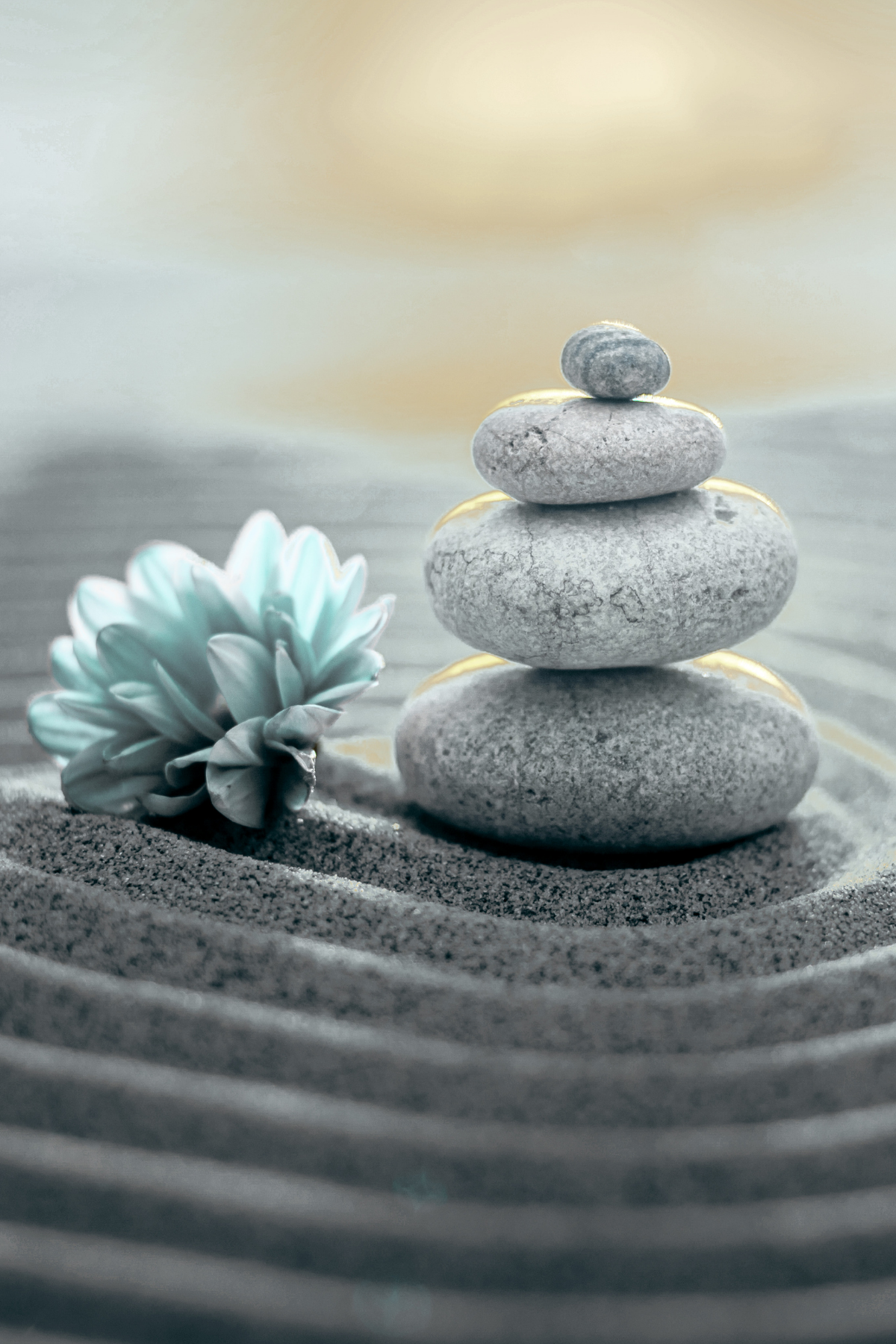 A stack of five smooth, gray stones balanced on top of each other in a sandy area, with a small flower nearby, and ripples in the sand surrounding them. The background shows a soft, warm light.