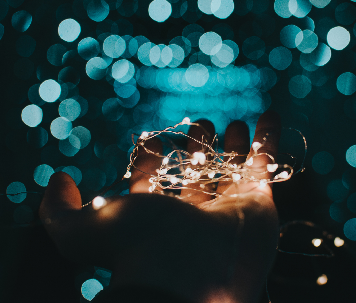 A hand holding a string of small, glowing fairy lights with a bokeh background of blue and teal lights.