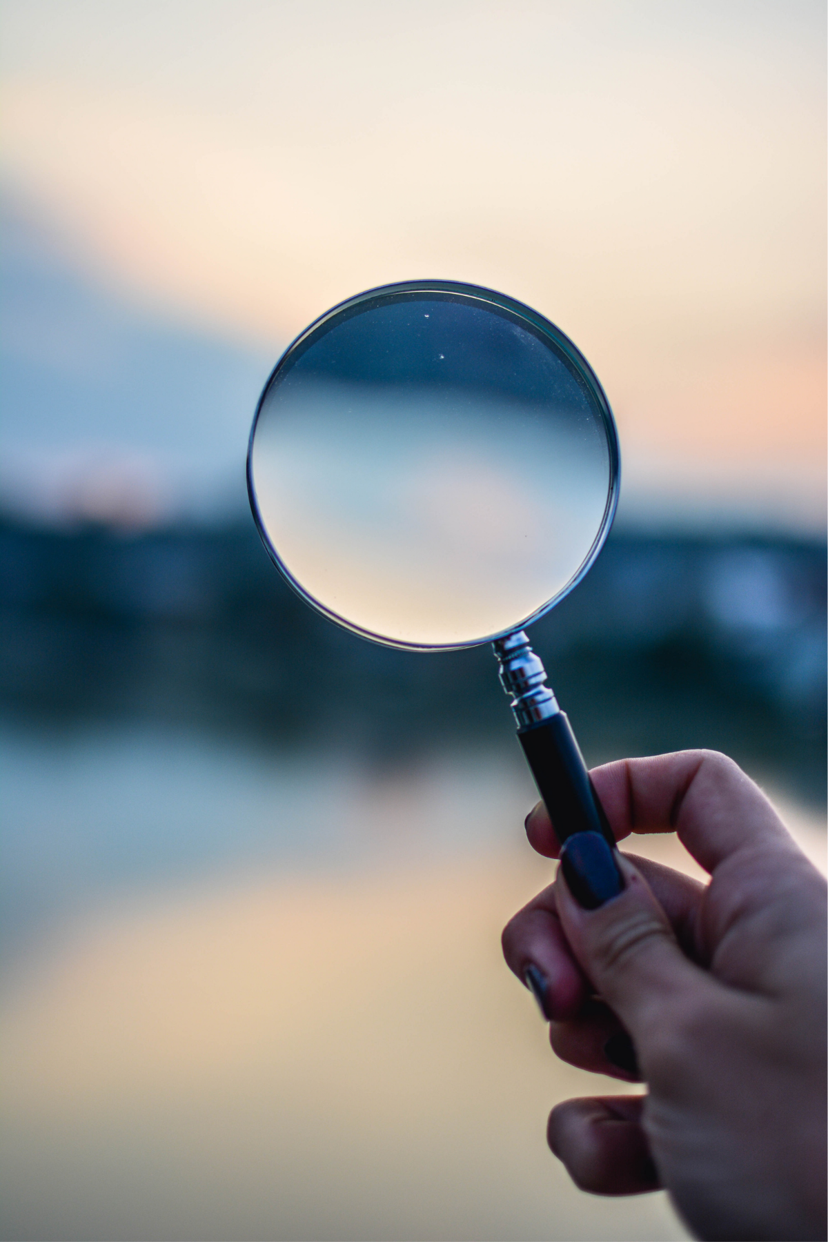 Close-up of a hand holding a magnifying glass against a blurred outdoor background during sunset or sunrise.