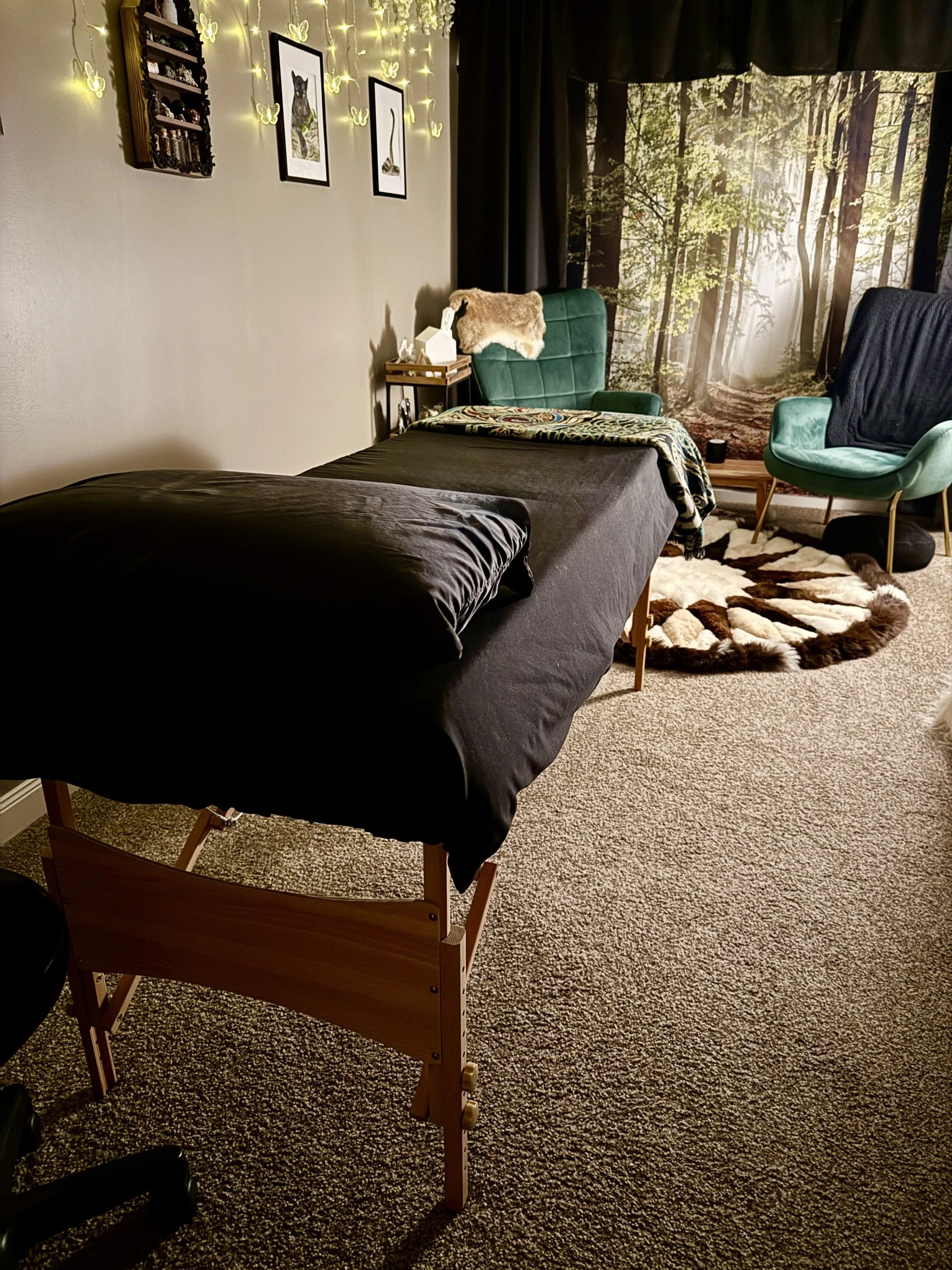 A room with a massage or therapy table covered with a black sheet, a walking cane, and a cozy seating area with a green chair and a teal armchair near a forest-themed wall mural; decorated with string lights, framed artwork, and butterfly decorations.