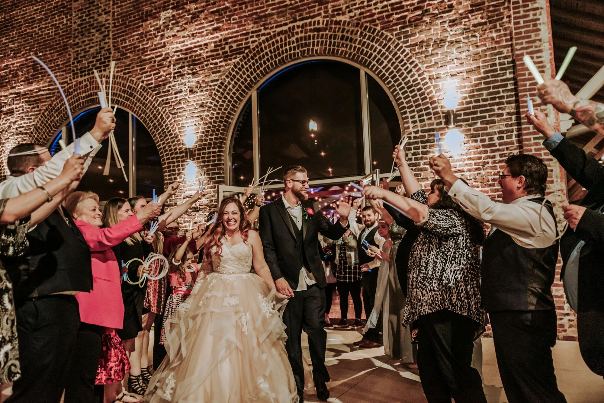 A newlywed couple walking hand-in-hand through a celebration with guests holding sparklers. The bride wears a lace wedding gown, and the groom is dressed in a black suit with a boutonniere. The setting is a brick-walled venue with warm lighting.