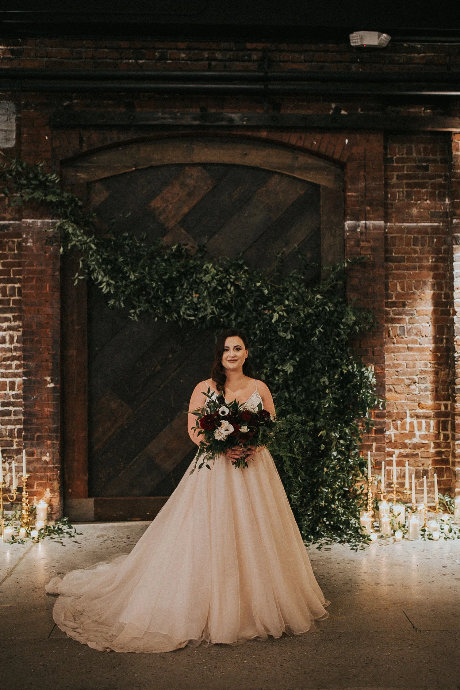 A bride in a white wedding gown holding a bouquet of flowers, standing indoors against a rustic brick wall with green foliage and candles.