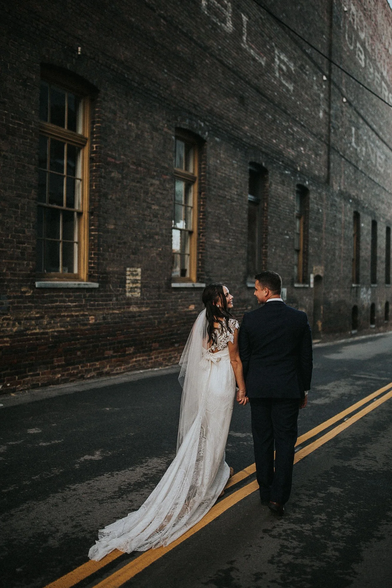 A newlywed couple walking through Downtown Knoxville at dusk, holding hands and smiling at each other, with a brick building in the background.