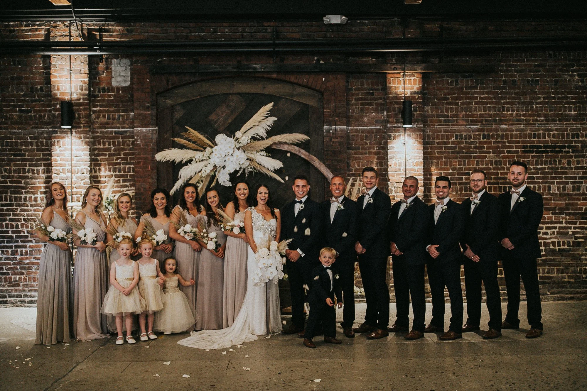 A wedding party standing in front of a brick wall with a floral arrangement backdrop, including the bride and groom, bridesmaids, groomsmen, flower girls, and ring bearer.