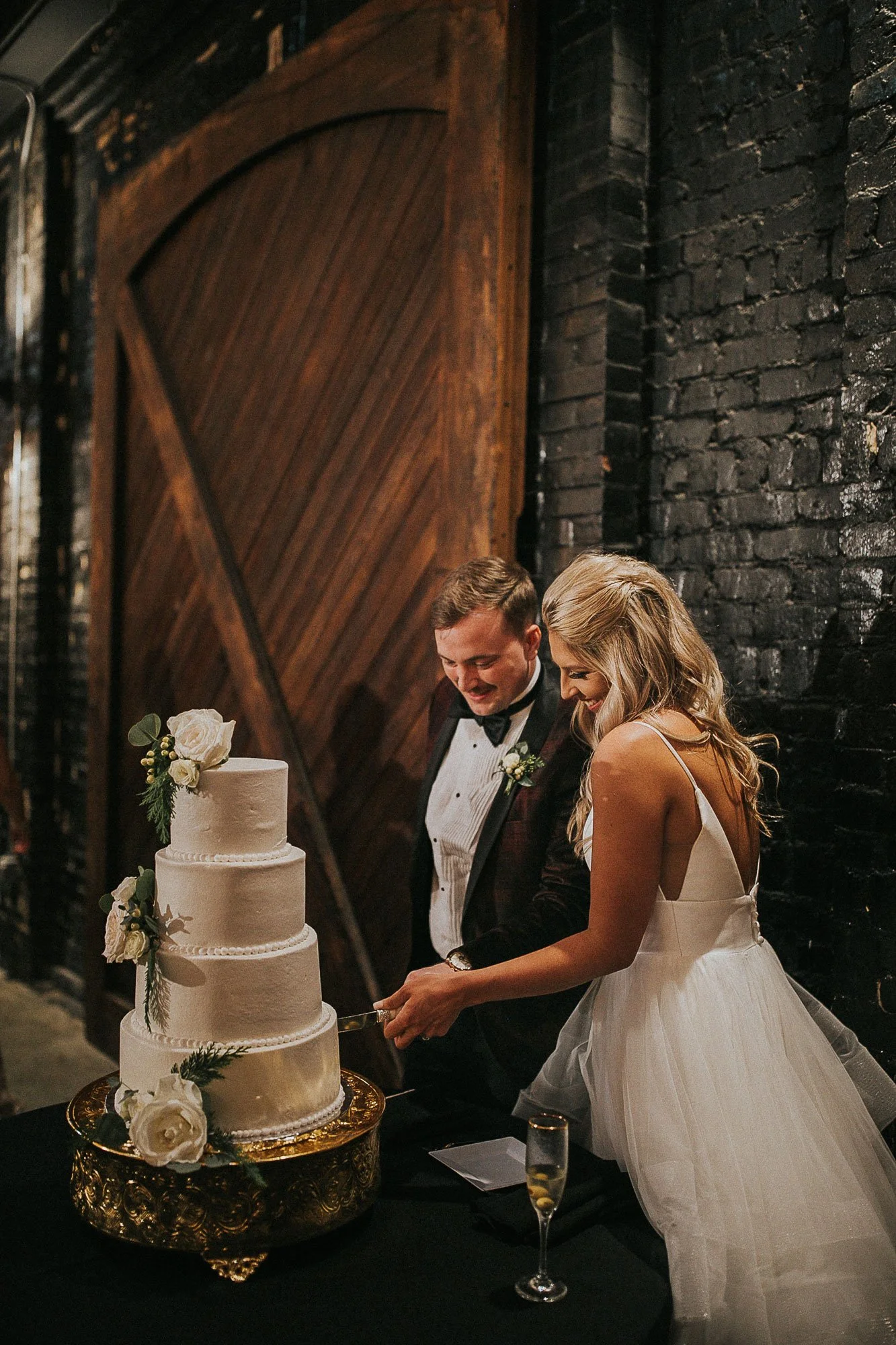 Bride and groom cutting a three-tier wedding cake decorated with white flowers and greenery at a wedding reception.