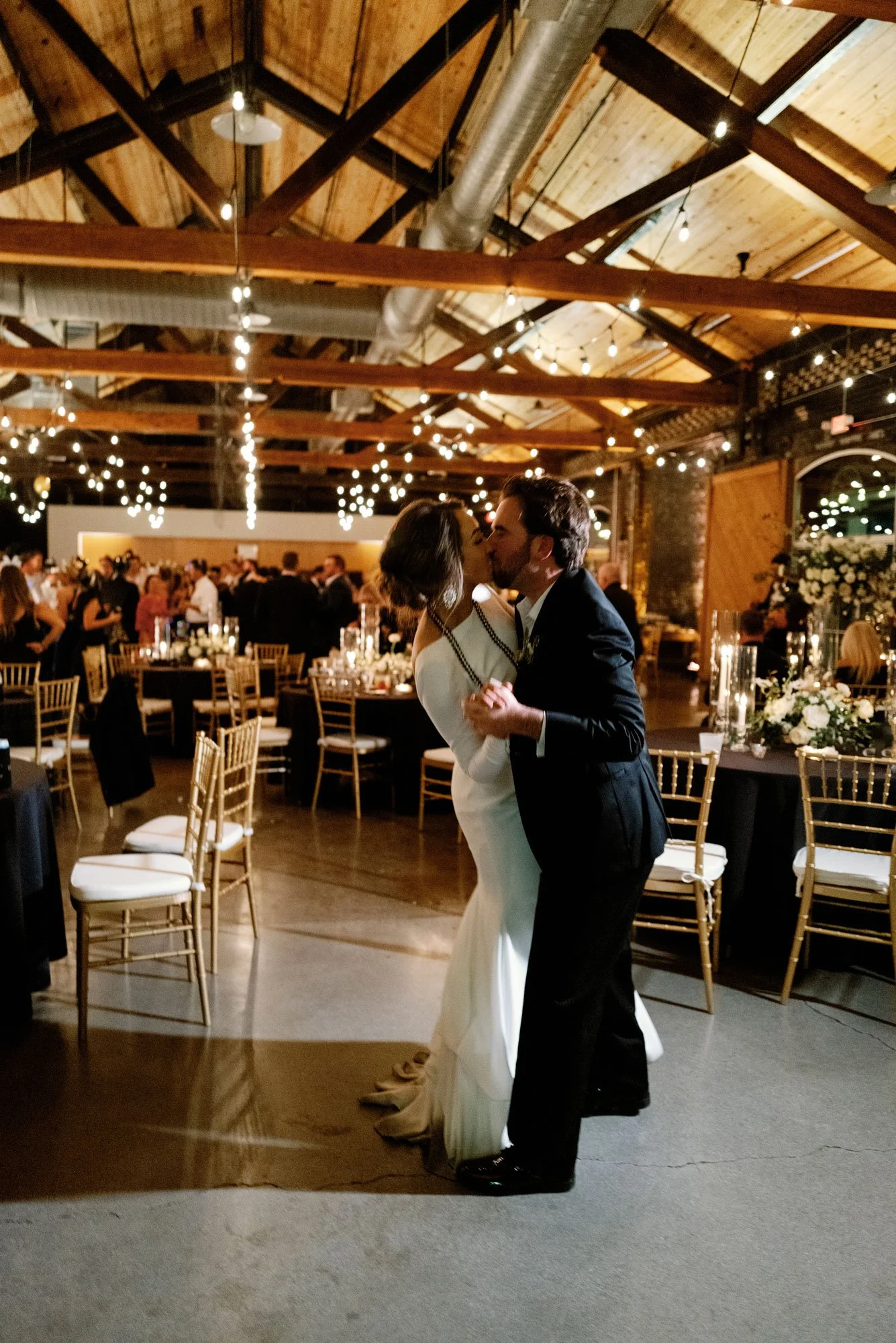 A newlywed couple kissing and dancing at their wedding reception in a decorated rustic venue with string lights and floral arrangements.