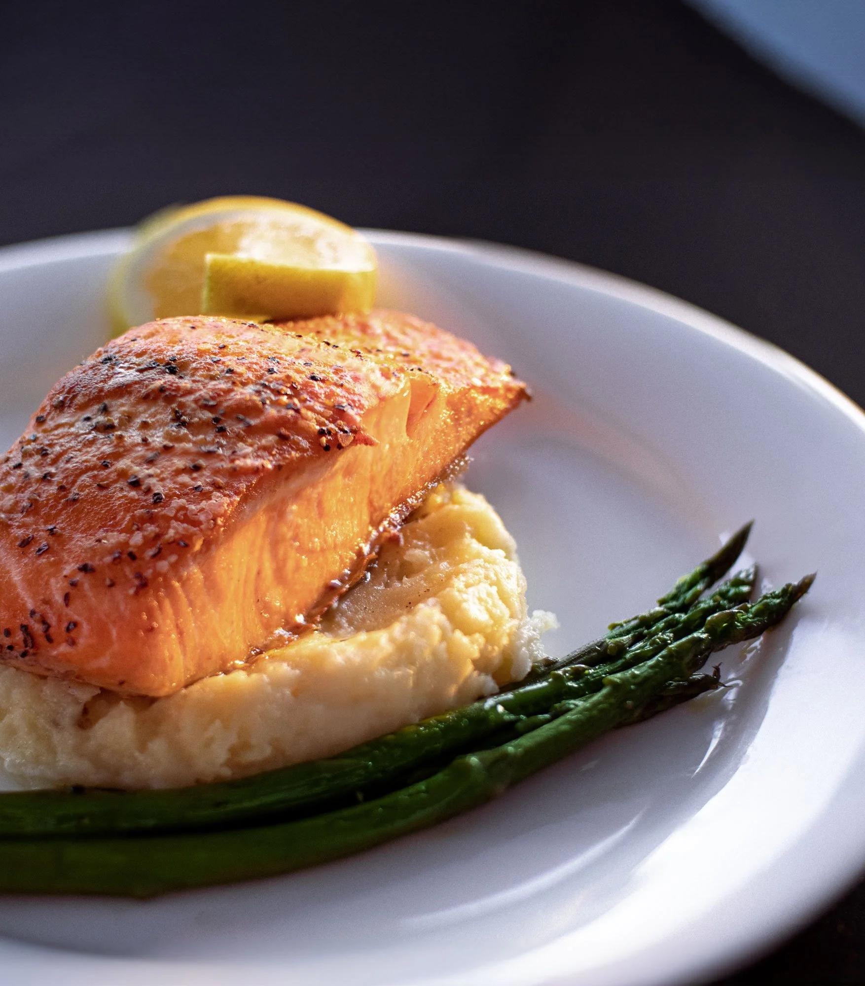 Pan-seared salmon fillet served on mashed potatoes with grilled asparagus and lemon wedge on a white plate.