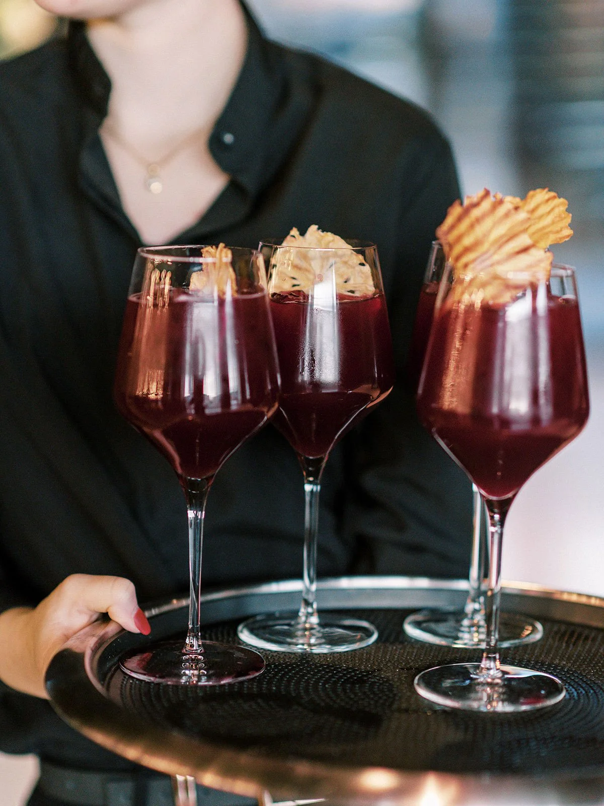 Person in black shirt holding a tray with three glasses of red wine garnished with potato chips.