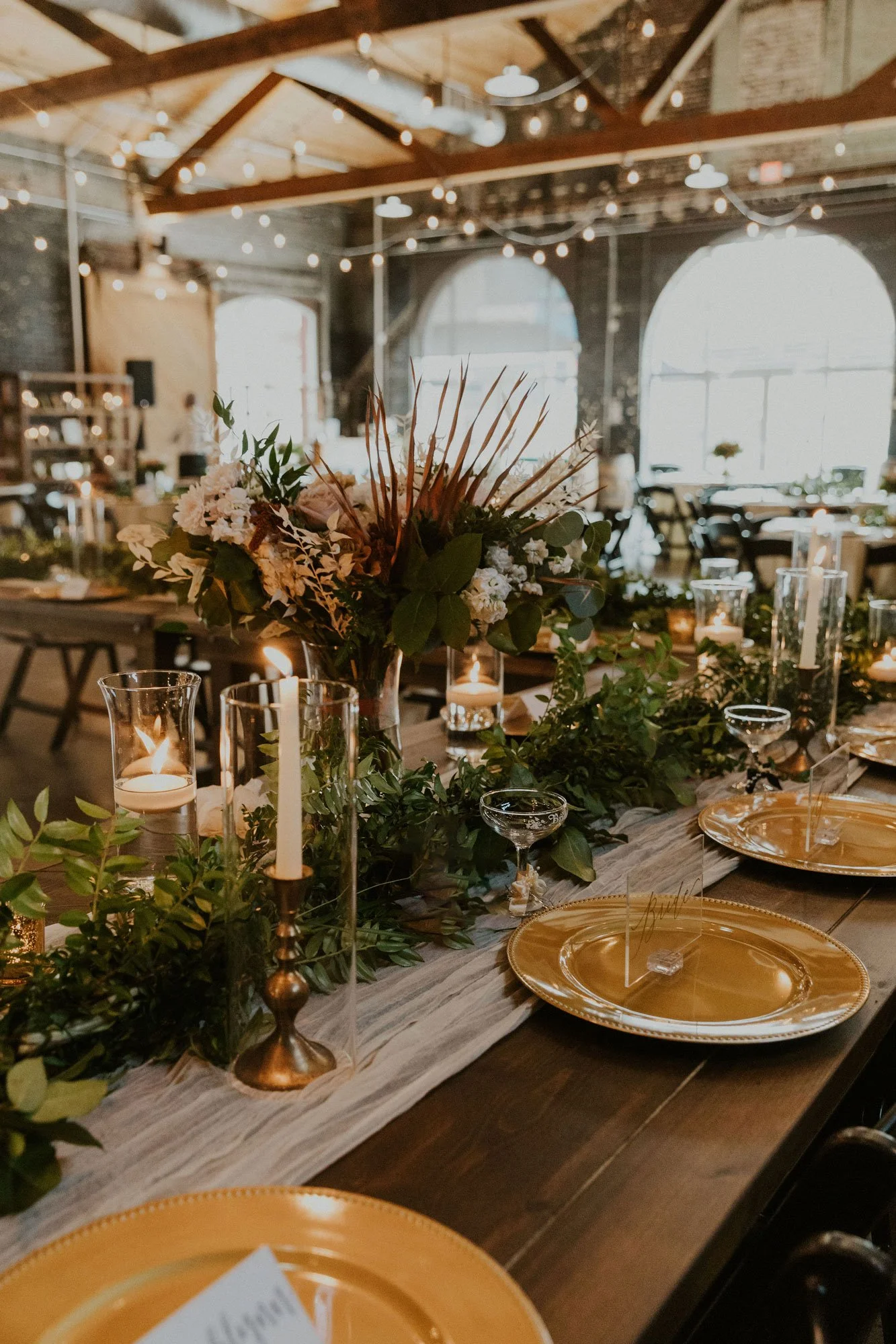 A decorated dinner table with gold plates, candles in glass holders, and a floral centerpiece in a rustic industrial venue with string lights and large windows.