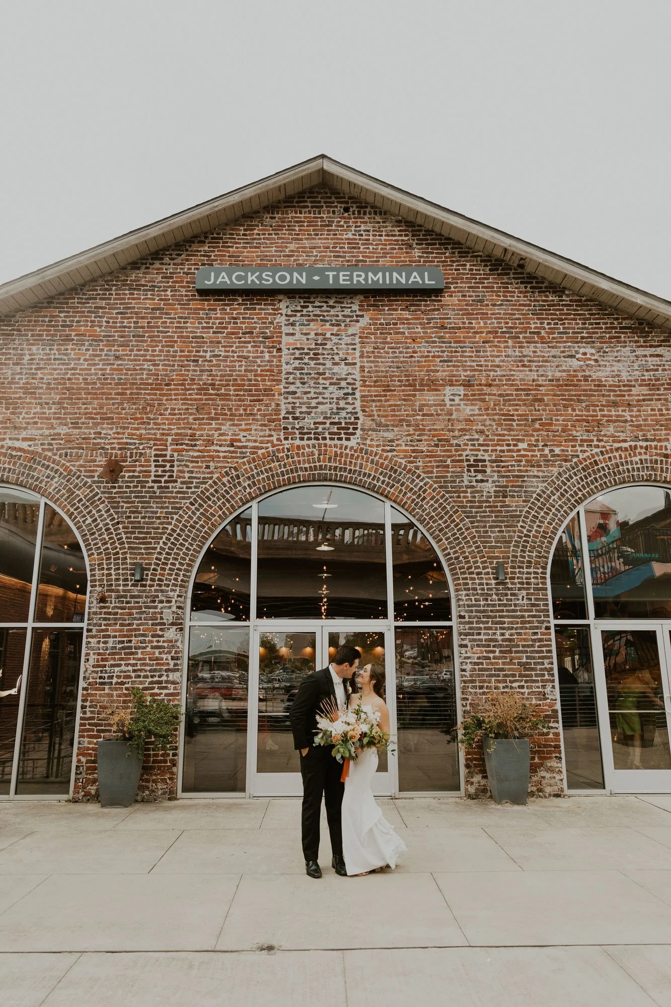 A bride and groom stand close together outside the Jackson Terminal building, holding a bouquet of flowers, with the building's brick facade and arched windows in the background.