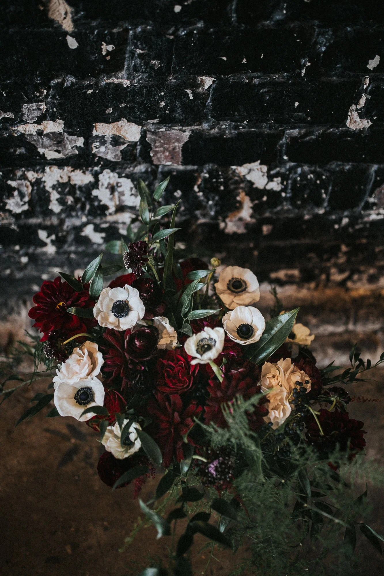 A bouquet of dark red, white, and purple flowers with green leaves placed against a black brick wall.