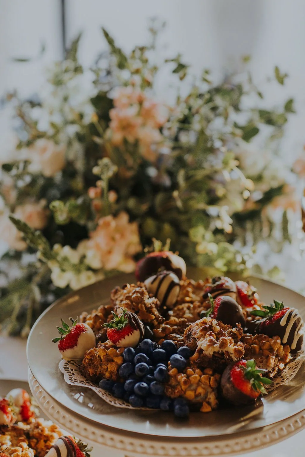 A dessert platter with strawberries, blueberries, and chocolate-covered snacks beside a floral arrangement.